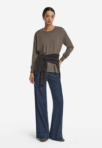 Brązowa bluzka z długim rękawem i związanym ciemnym paskiem w talii, połączona z szerokimi spodniami dżinsowymi w ciemnym kolorze. Stylizacja charakteryzuje się luźnym krojem i prążkowanymi mankietami.