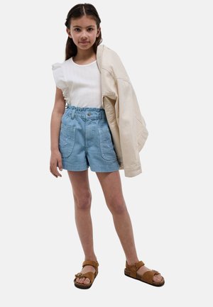 Jeune fille portant un haut blanc à manches volantées, un short bleu taille haute avec des poches, des sandales marron, et tenant une veste beige sur une épaule.