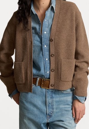 Femme portant un cardigan en maille marron par-dessus une chemise en jean bleue boutonnée, rentrée dans un jean bleu taille haute avec une ceinture marron.