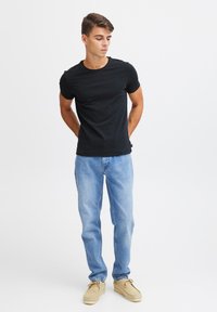 Maglietta nera a maniche corte abbinata a jeans azzurri chiari e mocassini beige in stile mocassino. Modello in piedi con le mani dietro la schiena su uno sfondo semplice.