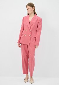 Vaaleanpunainen kaksirivinen bleiseri, jossa on kultaiset napit, väljästi istuvat housut ja beigellä korkokengät. Sujuva pinta, räätälöity malli.