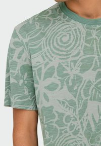 Camiseta de manga corta de color verde claro, hecha de una tela suave, que presenta un gran patrón floral blanco y en espiral en la parte delantera.
