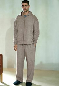 Brun fleece hættejakke med lynlås og hætte, kombineret med matchende bukser i afslappet pasform. Glat tekstur, to sidelommer og enkelt design.