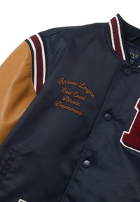 Marineblauwe varsityjack met bruine leren mouwen, rood-witte gestreepte kraag, geborduurde tekst en een grote rode letter op de voorkant.