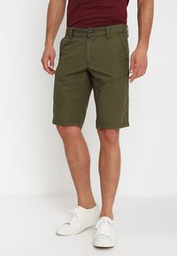 Shorts vert olive en mélange de coton, avec une coupe décontractée, des poches latérales et des coutures angulaires, associés à des baskets blanches.