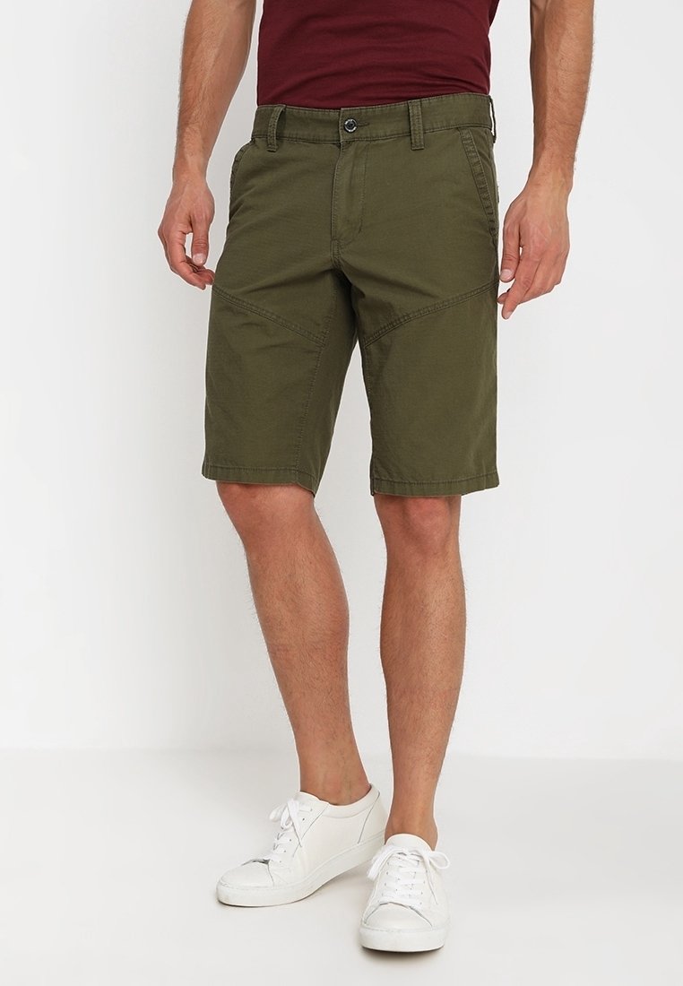 Shorts vert olive en mélange de coton, avec une coupe décontractée, des poches latérales et des coutures angulaires, associés à des baskets blanches.