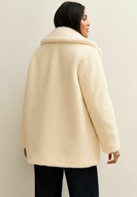 Cappotto in pelliccia sintetica color crema con un ampio colletto, vestibilità over size e maniche lunghe, caratterizzato da una superficie morbida e testurizzata.