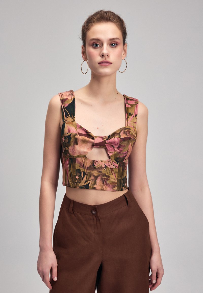 Crop top med levende blomsterprint i pink og grøn, med brede stropper og en sløjfedetalje foran. Brune højtaljede bukser.
