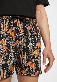 Shorts noirs avec un motif floral orange et blanc, ceinture élastique avec un cordon de serrage noir, portés avec un t-shirt noir ; main détendue sur le côté.