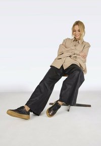 Beige linen button-up shirt, black leather wide-leg pants, and black sneakers with gum soles, seated on a stool against a light backdrop.