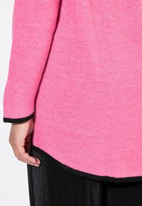 Fuchsia stickad tröja med rundad fåll och svart kant, bärs över veckad svart tyg. Mjuk textur med synliga stickningsmönster.