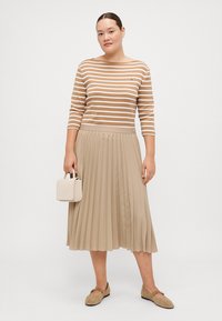 Beige Faltenrock kombiniert mit einem gestreiften braun-weißen Oberteil, trägt eine kleine cremefarbene Handtasche und beige Schuhe.