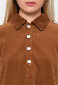 Femme portant une chemise en velours côtelé marron avec un col et quatre boutons-pression en argent visibles sur le devant.