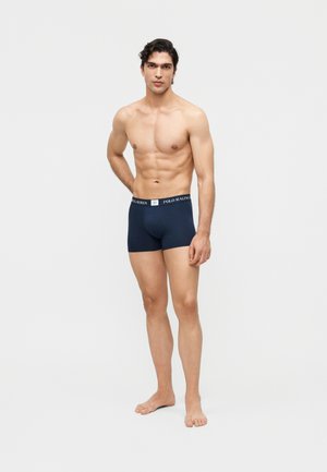 Paita päällä seisova mies paljain jaloin, pukeutuneena tummansinisiin Polo Ralph Lauren -bokserialushousuihin, valkoisen yksivärisen taustan edessä.