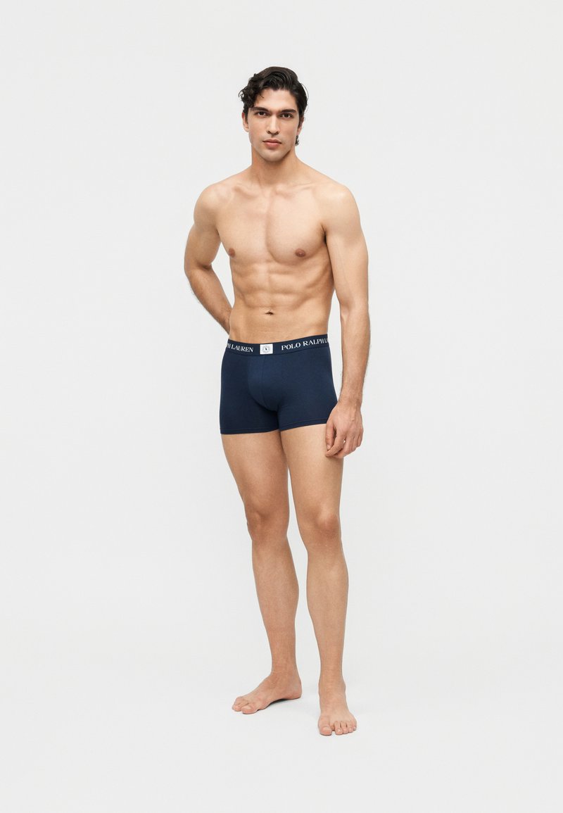 Bărbat fără tricou, stând desculț, purtând chiloți boxer Polo Ralph Lauren bleu închis, pe un fundal alb simplu.