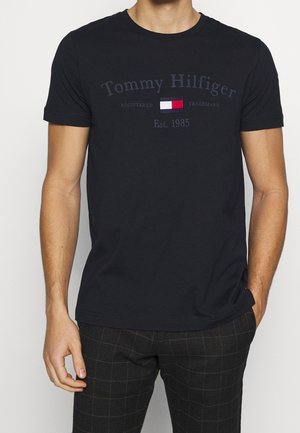 Mężczyzna w czarnej koszulce Tommy Hilfiger z logo i napisem "Est. 1985," w połączeniu z ciemnymi spodniami w kratę, stoi z jedną ręką w kieszeni.
