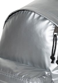 Zaino grigio con un tessuto liscio e lucido. Presenta una parte superiore arrotondata, una tasca frontale e hardware del cordoncino nero. Design pulito senza motivi.