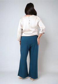 Lichtbeige satijnen top met driekwart mouwen en een knoopdetail op de rug, gecombineerd met teal wijde broek met zijsplitten.