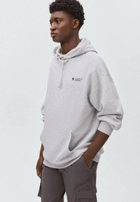 Hoodie gris avec une poche avant et une capuche à cordons, arborant un petit logo noir avec l'inscription "CITOYEN DE NULLE PART" sur la poitrine. Coupe décontractée.