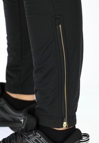 Black athletic pants with a zipper on the lower leg, featuring gold hardware. They have a fitted design and smooth texture. Black sneakers are visible.