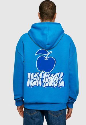 Felpa blu con una grande grafica di mela bianca e testo stilizzato "NEW YORK" sul retro. Il materiale sembra morbido e ha una vestibilità comoda.