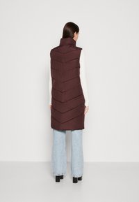 Gilet matelassé bordeaux avec un col haut, présentant un motif en chevrons et une texture lisse, associé à un jean large délavé.