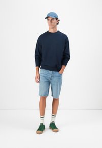 Navy sweater, denim shorts, groene sneakers, gestreepte sokken. Casual look met een lichtblauwe pet, met een effen patroon en een relaxte pasvorm.