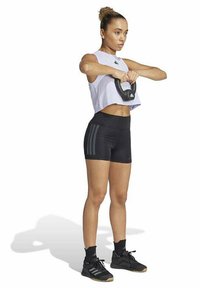 Sportieve crop top in lichtpaars, zwarte shorts met drie horizontale strepen en zwarte sneakers; houdend een kettlebell, geconcentreerde houding.