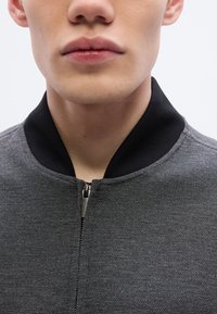 Γκρι ζακέτα με φερμουάρ, με μαύρο ριμπ γιακά, υφασμάτινη υφή και διακριτική γυαλάδα, με έμφαση σε κοντινό πλάνο του λαιμού.