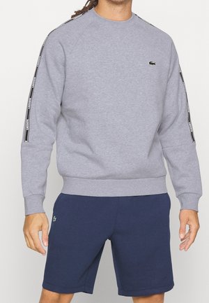 Mann trägt ein graues Sweatshirt mit schwarz-weißem Streifenlogo auf den Ärmeln und marineblaue Shorts mit kleinem Logo am linken Bein