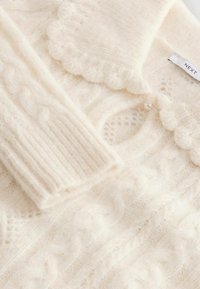 Cream knitted cardigan featuring a cable knit pattern, scalloped neckline, ribbed cuffs, and a pearl button accent.