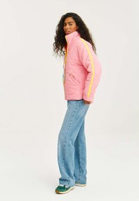 Pletená růžová bunda s žlutými pruhy po stranách, vysokým límcem, dvěma předními kapsami, nošená s světle modrými rozšířenými džíny a zelenými teniskami.