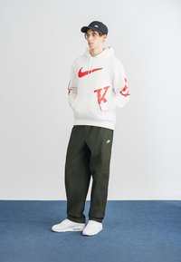 Sweatshirt branca com grandes logótipos vermelhos da Nike, combinada com calças de carga verde oliva. Ténis brancos completam o look. Boné preto na cabeça do modelo.