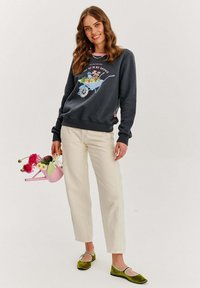 Tmavošedá mikina s grafikovou potlačou modrej kolečka naplneného kvetmi, krémové nohavice a zelené semišové topánky so svetlým lemovaním.