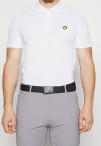 Valkoinen lyhythihainen polo-paita, jossa on keltainen perhoslogo, yhdistettynä harmaisiin housuihin ja mustaan vyöhön, jossa on suorakulmainen metallinen solki.