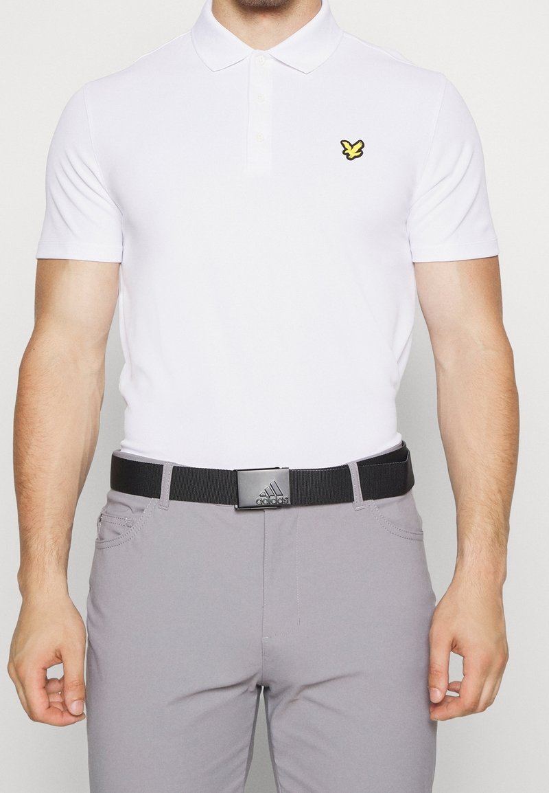 Valkoinen lyhythihainen polo-paita, jossa on keltainen perhoslogo, yhdistettynä harmaisiin housuihin ja mustaan vyöhön, jossa on suorakulmainen metallinen solki.