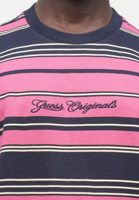 Maglietta a righe a maniche corte in rosa, blu navy e crema. Presenta collo a coste a girocollo e logo ricamato "Guess Originals" sul petto.