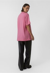 Roza oversized majica s kratkimi rokavi, okroglo izrezano vratno linijo in gladko teksturo, v kombinaciji z črnimi usnjenimi hlačami in obsežnimi črnimi čevlji.