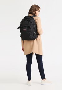 Mochila negra con múltiples compartimentos, correas ajustables y cremalleras. Llevada en la espalda por una persona con un abrigo beige y jeans oscuros.