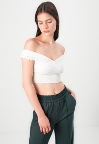 Petit top blanc à épaules dénudées avec des manches volantes, présentant un design à nouer à l'avant et un ourlet festonné, associé à un pantalon vert foncé à taille élastique.