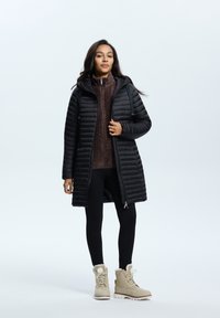 Schwarze Pufferjacke mit Kapuze, Reißverschluss und gerippter Textur. Kombiniert mit einem braunen Sweater und schwarzen Leggings, ergänzt durch beigefarbene Stiefel.