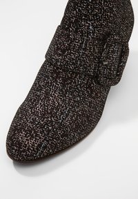 Botín negro con tejido texturizado que presenta un patrón moteado multicolor. Diseño de punta puntiaguda con un detalle de hebilla lateral.