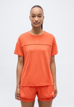 Femme portant un t-shirt de sport orange vif et un short assorti, debout face à l'avant sur un fond blanc uni.