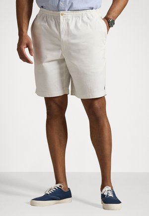 Homme portant un short beige clair, des baskets bleues et blanches, et une chemise bleue rayée rentrée, visible de la taille aux pieds.