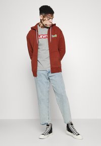 Sudadera de forro polar burdeos con cremallera y dos bolsillos frontales, camiseta gris con el logo rojo de Levi's, jeans de pierna recta azul claro y zapatillas negras de caña alta.