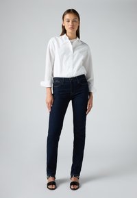 Camicia bianca a maniche lunghe con le maniche arrotolate, abbinata a jeans skinny blu scuro. Il modello indossa sandali neri con cinghie alla caviglia. Tessuti di stoffa lisci.