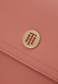 Vaaleanpunainen teksturoitu nahkalaukku, jossa on kultainen pyöreä logo, jossa on toisiinsa liittyvät kirjaimet, ja kontrastivärinen ommel reunan ympärillä.