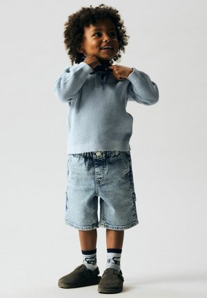 Jeune enfant aux cheveux bouclés portant un pull bleu, un short en jean, des chaussettes à motifs et des chaussures sombres à enfiler, ajustant une écharpe.