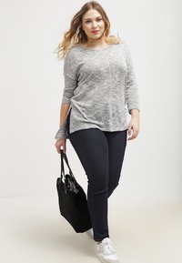 Svijetlosiva, dugosleeve pletena majica s bočnim prorezima u kombinaciji s tamnim trapericama i bijelim tenisicama, drži crnu torbu na rame. Mekana tekstura, ležeran kroj.