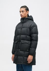 Giacca a vento nera con cappuccio, caratterizzata da una chiusura lampo frontale, grandi tasche anteriori e una texture trapuntata. Indossata sopra jeans blu.